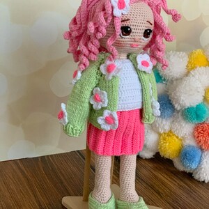 May include: A crocheted doll with pink curly hair, wearing a green cardigan with white flowers, a white shirt, a pink skirt, and green shoes. The doll is standing on a wooden star-shaped base.