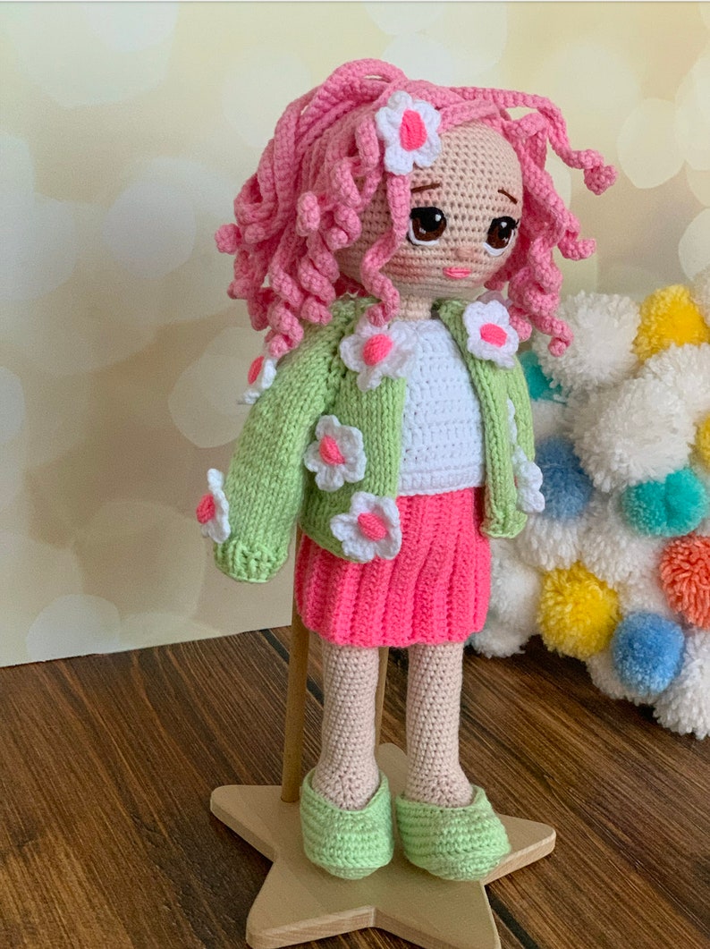 May include: A crocheted doll with pink curly hair, wearing a green cardigan with white flowers, a white shirt, a pink skirt, and green shoes. The doll is standing on a wooden star-shaped base.