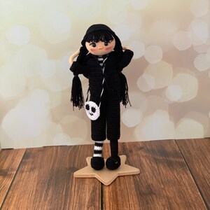 K&ouml;nnte beinhalten: Eine schwarz-wei&szlig;e H&auml;kelpuppe mit schwarzen Haaren, die ein schwarz-wei&szlig; gestreiftes Hemd, eine schwarze Hose und schwarz-wei&szlig; gestreifte Socken tr&auml;gt. Die Puppe h&auml;lt eine schwarz-wei&szlig;e H&auml;keltasche mit einem Totenkopfdesign.