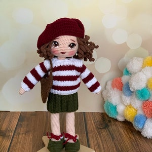 May include: A crocheted doll wearing a red and white striped jumper, a green skirt, a red beret, and brown shoes. The doll has brown curly hair and is standing on a wooden star.