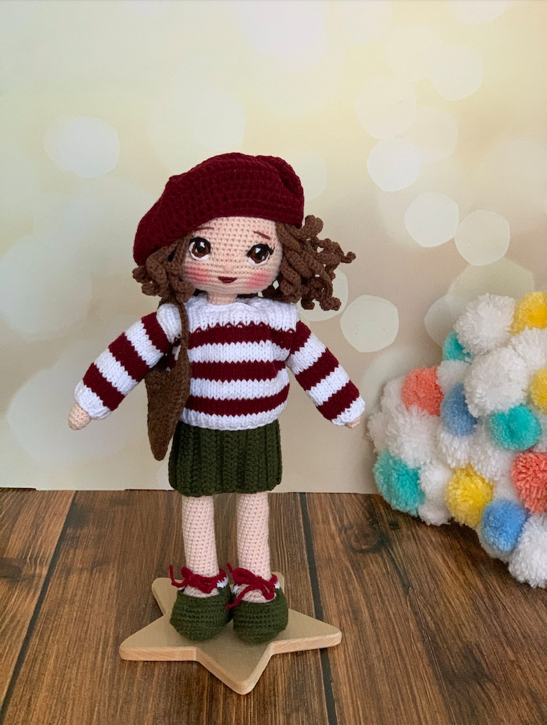 May include: A crocheted doll wearing a red and white striped jumper, a green skirt, a red beret, and brown shoes. The doll has brown curly hair and is standing on a wooden star.