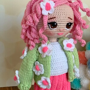 May include: A crocheted doll with pink curly hair, a white shirt, a green cardigan with white flower accents, and a pink skirt. The doll has brown eyes and a pink flower in her hair.