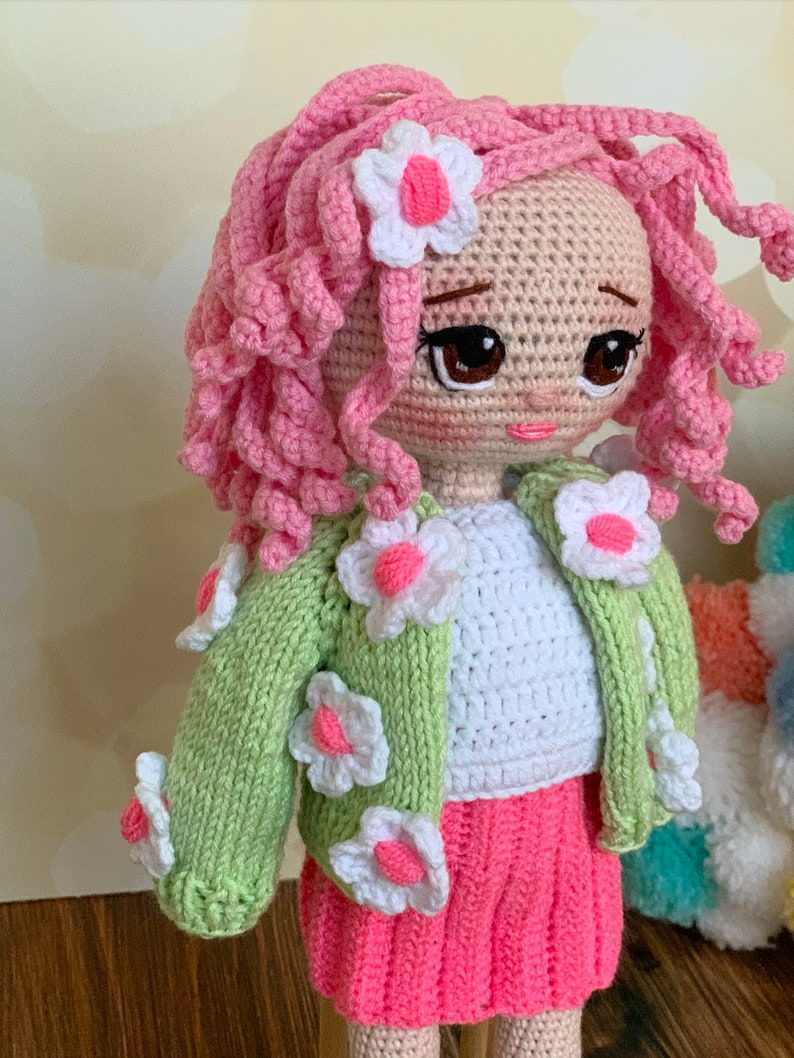 May include: A crocheted doll with pink curly hair, a white shirt, a green cardigan with white flower accents, and a pink skirt. The doll has brown eyes and a pink flower in her hair.