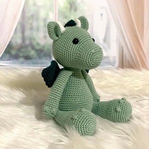 May include: A green crocheted dragon plush toy with a dark green wing and white accents on the head. The dragon is sitting on a white fluffy surface.