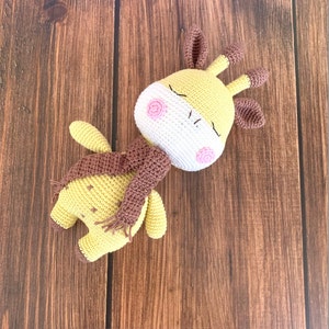 May include: A crocheted giraffe toy with a brown scarf, wearing a white face mask with pink accents. The toy is lying on a wooden surface.