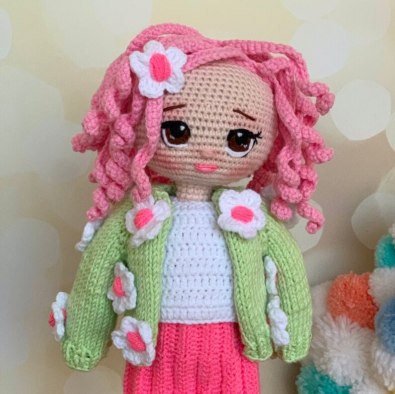 May include: A crocheted doll with pink curly hair, wearing a green cardigan with white flowers, a white shirt, and a pink skirt.