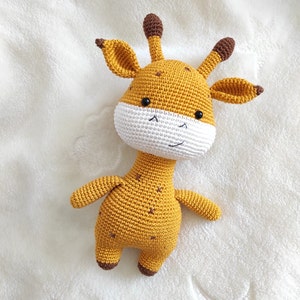May include: A crocheted giraffe toy with a white face and brown spots. The giraffe is lying on a white fluffy surface.