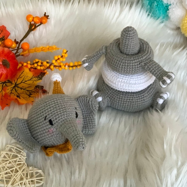 May include: Two grey crocheted elephant toys. One elephant has a party hat and bow tie. The other is a stackable toy with a white stripe. The toys are on a white fluffy surface.