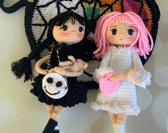 Könnte beinhalten: Zwei gehäkelte Puppen, eine mit schwarzen Haaren und einem schwarz-weißen Kleid, die andere mit rosa Haaren und einem weißen Kleid. Beide Puppen halten kleine gehäkelte Handtaschen. Die Puppen sitzen vor einer gehäkelten Tasche mit einem Regenbogen- und Spinnennetz-Design.