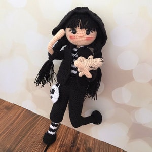 K&ouml;nnte beinhalten: Eine geh&auml;kelte Puppe mit schwarzen Haaren, die ein schwarz-wei&szlig; gestreiftes Shirt, eine schwarze Hose und eine schwarze Kapuzenjacke tr&auml;gt. Die Puppe h&auml;lt ein geh&auml;keltes Stofftier.