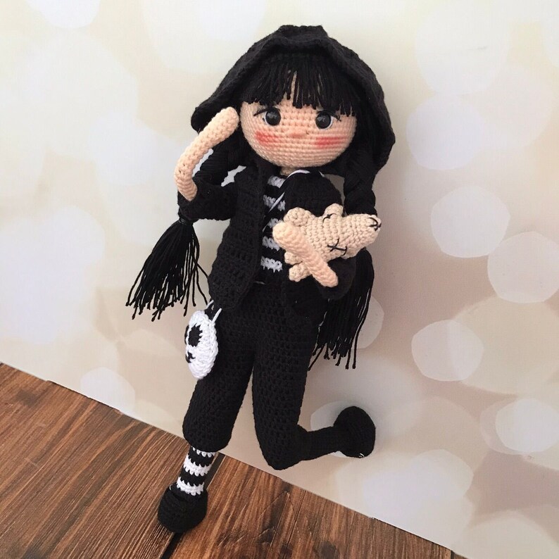 K&ouml;nnte beinhalten: Eine geh&auml;kelte Puppe mit schwarzen Haaren, die ein schwarz-wei&szlig; gestreiftes Shirt, eine schwarze Hose und eine schwarze Kapuzenjacke tr&auml;gt. Die Puppe h&auml;lt ein geh&auml;keltes Stofftier.