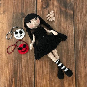 May include: Crocheted doll with black hair, a black dress, and white and black striped socks. The doll is holding a black and white crocheted bag with a skull and crossbones design. There are two other crocheted bags, one red with a smiling face and one white with a skull and crossbones design.
