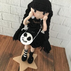 May include: A crocheted doll with black hair, a black dress, and a white skull-shaped purse. The doll is wearing black and white striped socks and black shoes. The doll is standing on a wooden star.