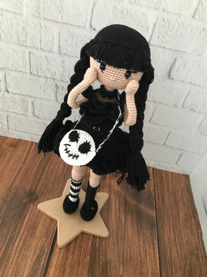 May include: A crocheted doll with black hair, a black dress, and a white skull-shaped purse. The doll is wearing black and white striped socks and black shoes. The doll is standing on a wooden star.