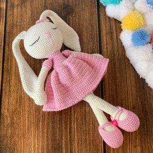 Puede incluir: Una muñeca de conejo de crochet blanco con un vestido rosa y zapatos rosas. La muñeca tiene los ojos cerrados y un lazo rosa en la cabeza.