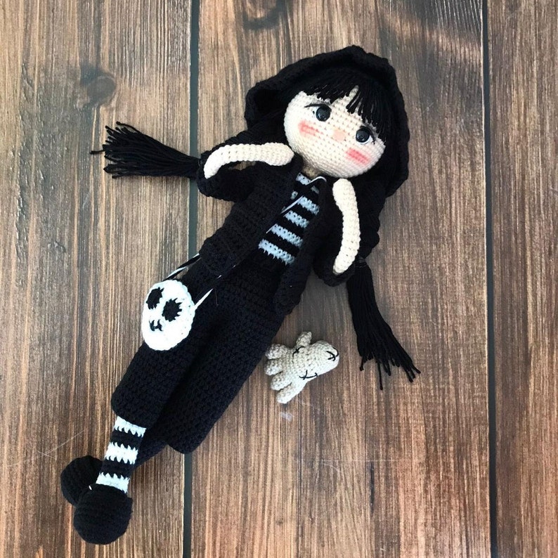 K&ouml;nnte beinhalten: Eine schwarze H&auml;kelpuppe mit einer wei&szlig;en Totenkopf-Handtasche, einem schwarz-wei&szlig; gestreiften Shirt und einer schwarzen Kapuzenjacke. Die Puppe hat schwarze Haare, schwarze Augen und rosa Wangen. Die Puppe h&auml;lt eine kleine wei&szlig;e H&auml;kelhand.
