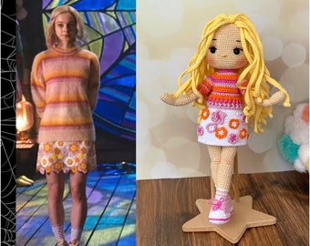Könnte beinhalten: Eine gehäkelte Puppe mit blonden Haaren, die ein pink-orangenes gestreiftes Shirt, einen weißen Rock mit pink-orangenen Blumen und weiße Sneaker trägt. Die Puppe steht auf einem sternförmigen Holzsockel.