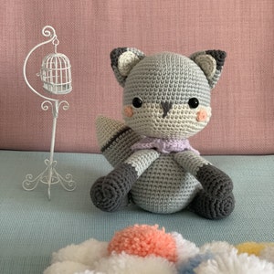 Puede incluir: Un juguete de peluche de zorro gris tejido a crochet con una bufanda morada. El zorro tiene ojos negros y mejillas rosadas. El juguete está sentado sobre una superficie azul claro.