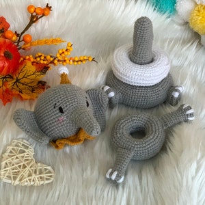 May include: Crocheted grey elephant toy with a yellow collar and a party hat. A grey stacking toy with a white ring and a grey ring-shaped toy with feet are also present. The toys are on a white, fluffy surface.