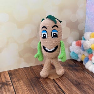 Op de afbeelding: Een gehaakt pluche speelgoed in de vorm van een glimlachende bruine aardappel met groene armen en benen. Het speelgoed heeft blauwe ogen en een witte glimlach.