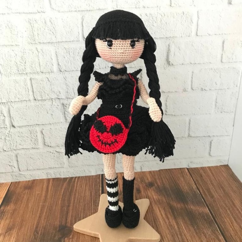 May include: Crocheted doll with black hair, a black dress, and black and white striped socks. The doll is holding a red purse with a black and white jack-o-lantern face.