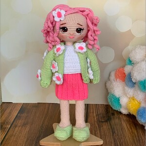 May include: A crocheted doll with pink curly hair, wearing a green cardigan with white flowers, a white shirt, a pink skirt, and green shoes. The doll is standing on a wooden star.