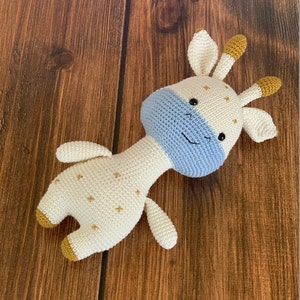 May include: A crocheted stuffed giraffe toy with a white body, blue face, and gold accents. The giraffe has a smiling expression and is lying on a wooden surface.