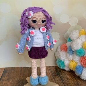 May include: A crocheted doll with purple hair, a blue cardigan with white flowers, a white shirt, a purple skirt, and blue shoes. The doll is standing on a wooden star.