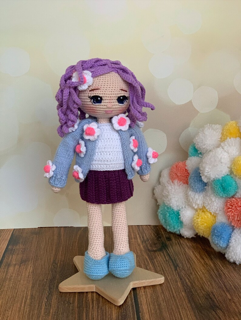 May include: A crocheted doll with purple hair, a blue cardigan with white flowers, a white shirt, a purple skirt, and blue shoes. The doll is standing on a wooden star.