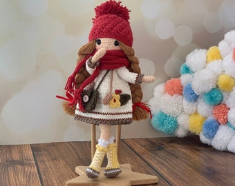 May include: A crocheted doll wearing a red hat, white dress, and brown boots. The doll has brown hair and is holding a brown bag. The doll is standing on a wooden star-shaped stand.