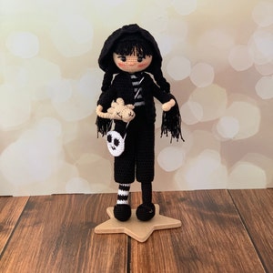 K&ouml;nnte beinhalten: Eine geh&auml;kelte Puppe, die eine schwarze Kapuzenjacke, eine schwarze Hose und schwarz-wei&szlig; gestreifte Socken tr&auml;gt. Die Puppe h&auml;lt ein kleines geh&auml;keltes Tier und hat eine Totenkopf-Tasche.