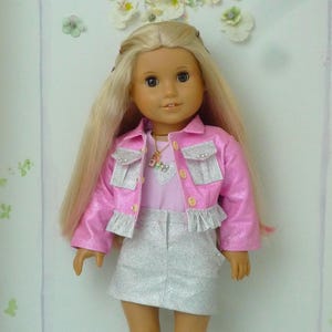 May include: A doll dressed in a pink jacket with silver detailing, a light pink top, and a silver skirt. The doll has long blonde hair and silver shoes. The background includes a floral decoration.