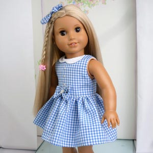 May include: A doll in a blue and white gingham dress with a white collar and a bow. The doll has long blonde hair styled with a braid and a blue bow. A pink hair clip is also visible.