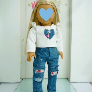 May include: A doll wearing a white long-sleeved shirt with a blue heart design and a gold necklace. The doll is wearing blue jeans with floral patches and has blonde hair.