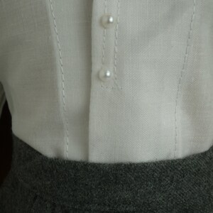 May include: Close-up of a white linen top with three pearl buttons and delicate stitching details. The top is paired with a gray, textured skirt. The image highlights the contrast between the textures and colors of the clothing.
