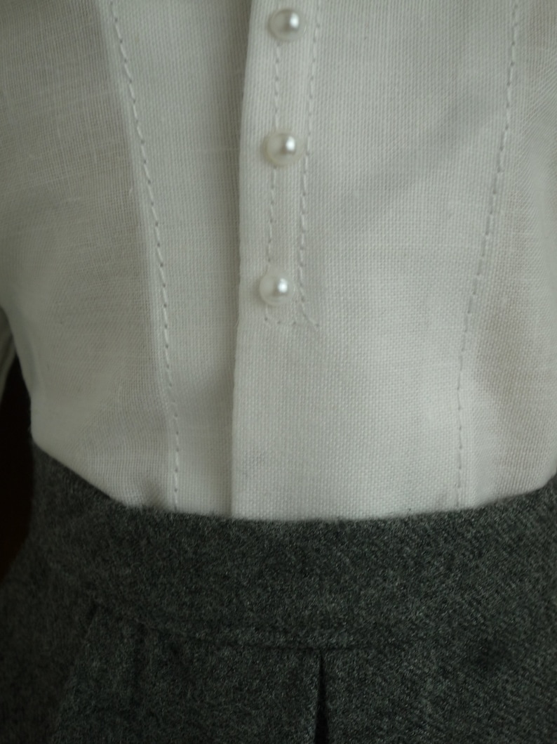 May include: Close-up of a white linen top with three pearl buttons and delicate stitching details. The top is paired with a gray, textured skirt. The image highlights the contrast between the textures and colors of the clothing.