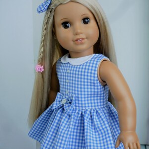May include: A doll with long blonde hair, styled with a blue gingham bow and a braid. The doll wears a blue and white gingham dress with a white collar and a bow at the waist. The dress is sleeveless.