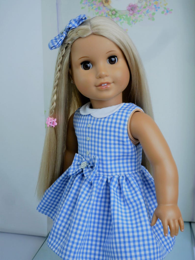 May include: A doll with long blonde hair, styled with a blue gingham bow and a braid. The doll wears a blue and white gingham dress with a white collar and a bow at the waist. The dress is sleeveless.