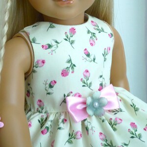 May include: A doll in a cream dress patterned with pink roses and green leaves. The dress has a pink bow with a light blue flower and pearl detail. The doll has blonde hair and a pink hair clip.