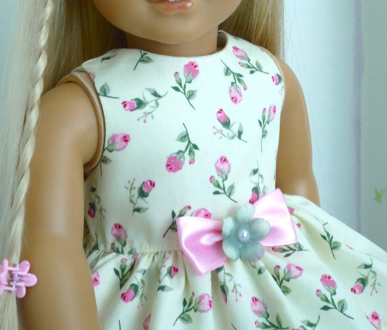 May include: A doll in a cream dress patterned with pink roses and green leaves. The dress has a pink bow with a light blue flower and pearl detail. The doll has blonde hair and a pink hair clip.