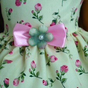 May include: A light yellow dress with a floral rose pattern in pink and green. The dress features a pink bow and a flower embellishment with a pearl centre. The dress is designed for a doll.