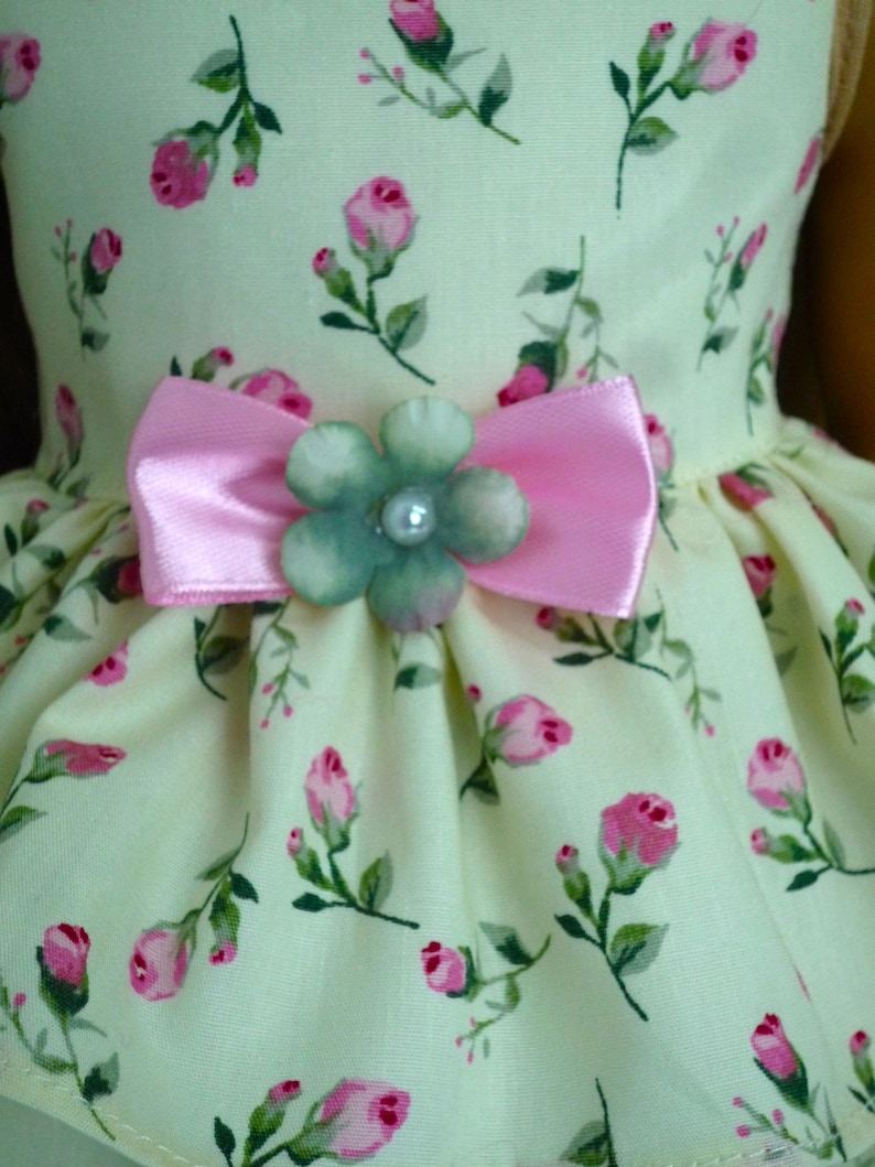 May include: A light yellow dress with a floral rose pattern in pink and green. The dress features a pink bow and a flower embellishment with a pearl centre. The dress is designed for a doll.