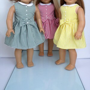 May include: Three dolls dressed in gingham dresses in green, pink, and yellow. Each dress features a white collar, a bow at the waist, and a sleeveless design. The dresses are made of a checkered fabric.