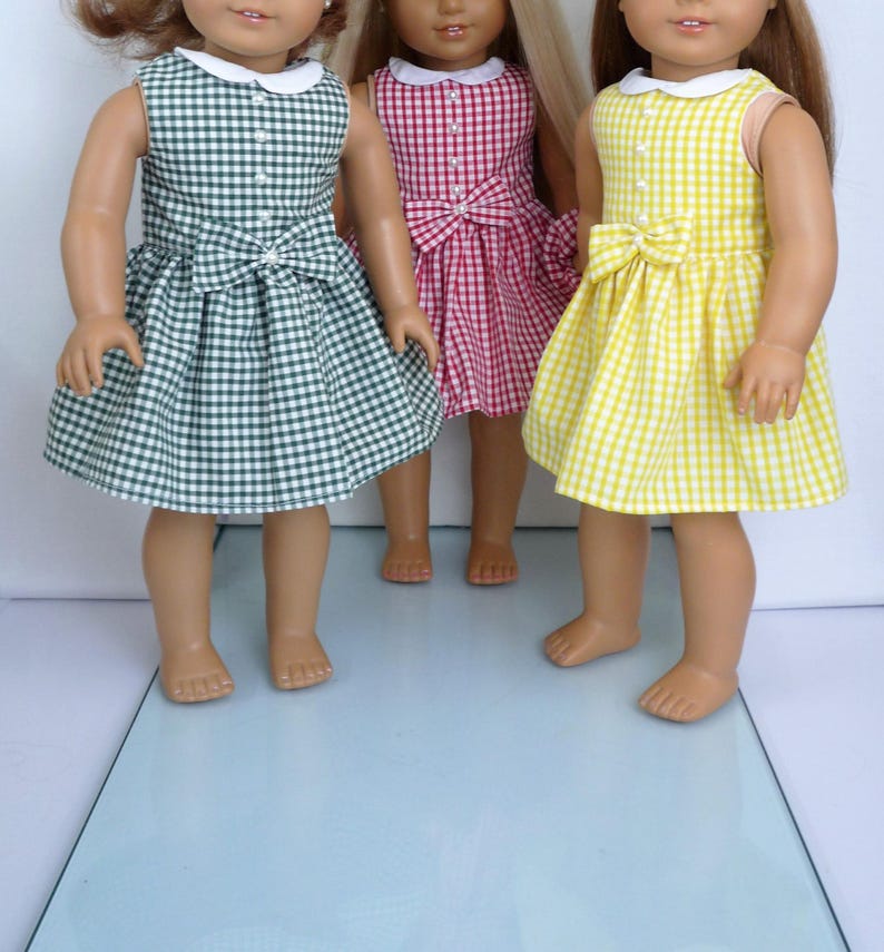 May include: Three dolls dressed in gingham dresses in green, pink, and yellow. Each dress features a white collar, a bow at the waist, and a sleeveless design. The dresses are made of a checkered fabric.