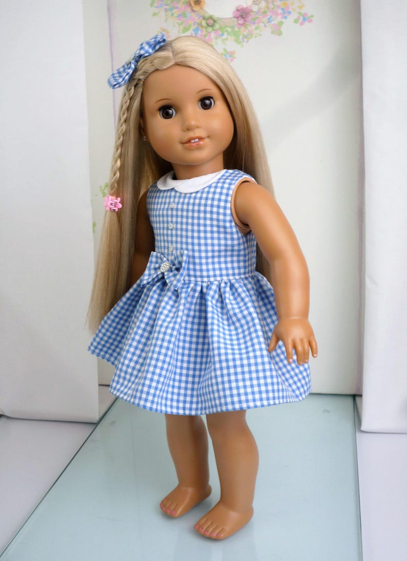 May include: A doll in a blue and white gingham dress with a white collar and a bow. The doll has long blonde hair styled with a braid and a blue bow. A pink hair clip is also visible.