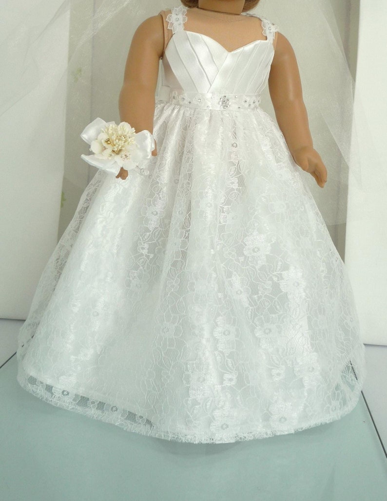 May include: White lace wedding dress with a satin bodice and a beaded belt. The dress has a full skirt and a lace trim. The doll is holding a white flower bouquet.