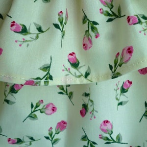 May include: Close-up of a cream fabric featuring a repeating pattern of pink roses and green leaves. The roses are in different stages of bloom, some as buds. The fabric looks like light cotton or linen.