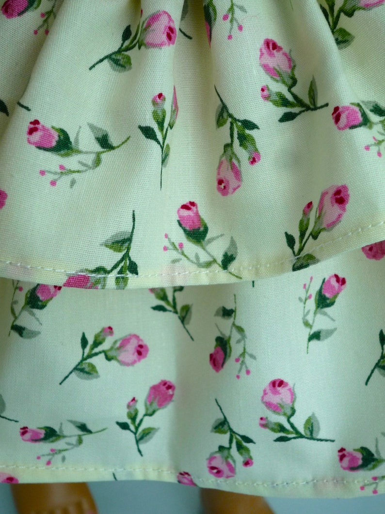 May include: Close-up of a cream fabric featuring a repeating pattern of pink roses and green leaves. The roses are in different stages of bloom, some as buds. The fabric looks like light cotton or linen.