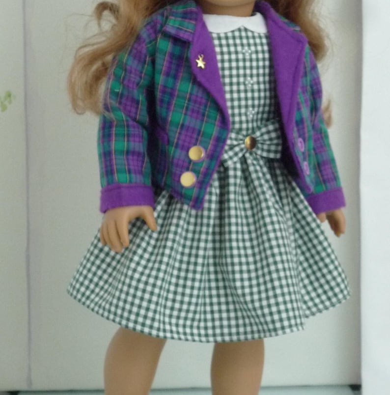 May include: A doll wearing a green and purple plaid jacket with a green and white gingham dress. The jacket has gold buttons and a star on the collar.
