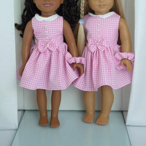 May include: Two dolls dressed in matching pink and white gingham dresses. The dresses feature white collars, pearl buttons, and a bow at the waist. Each doll also has a matching ruffled cuff. One doll has dark hair, and the other has blonde hair.